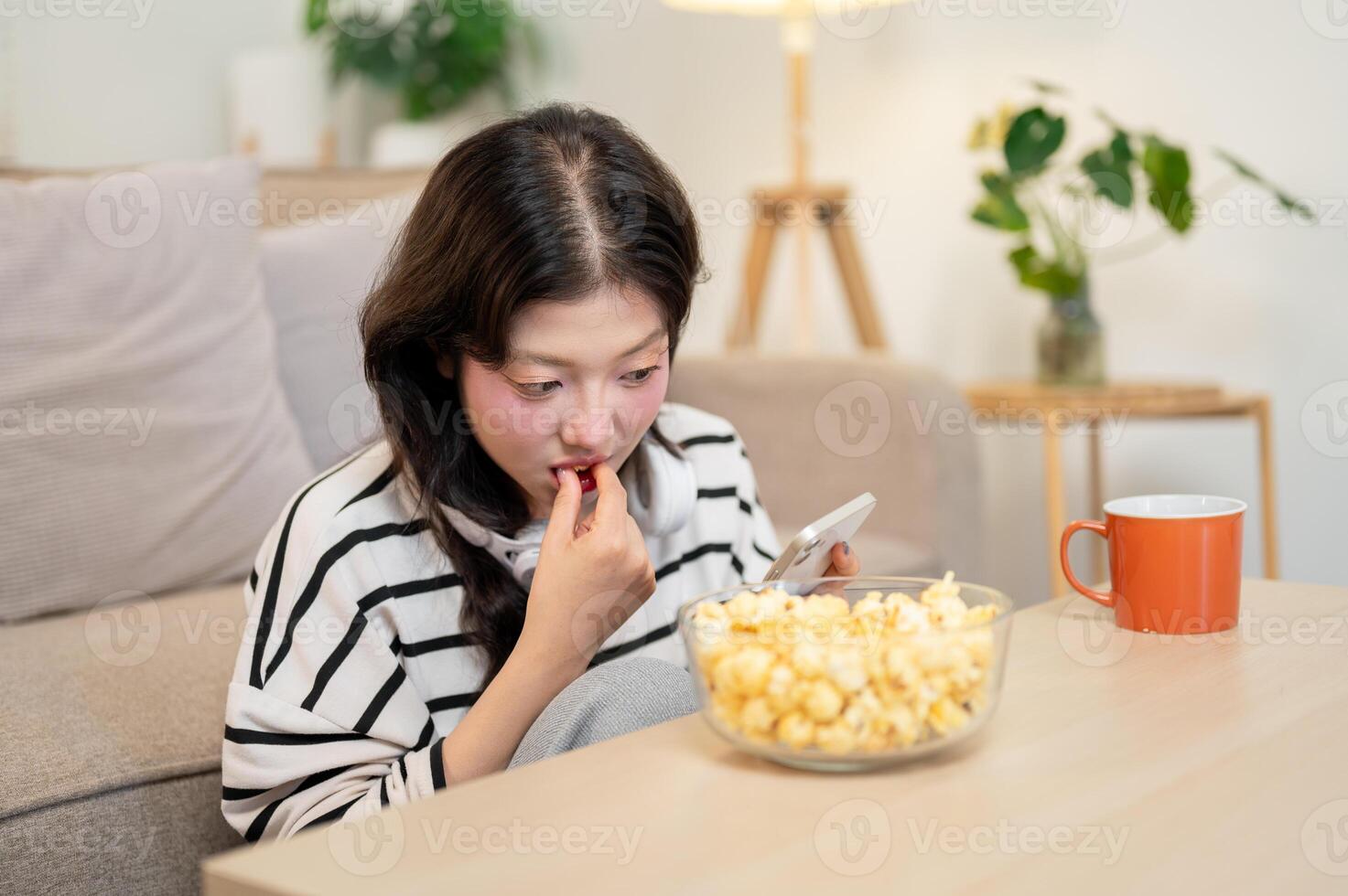 A relaxed woman is enjoying popcorn while reading chats on her phone, spending her free day at home. photo