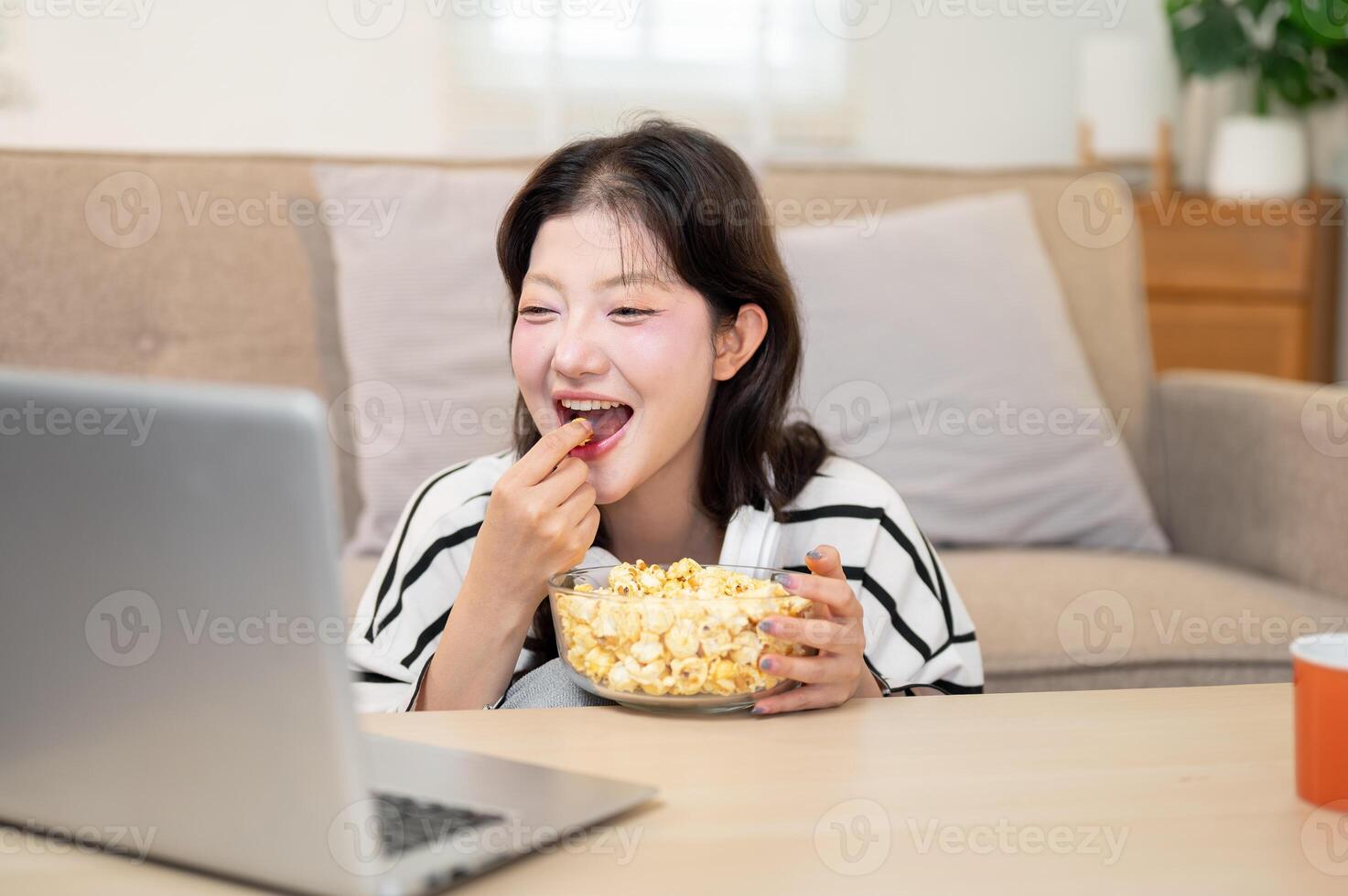 A cheerful Asian woman enjoys eating popcorn while watching a movie or game show on her laptop. photo