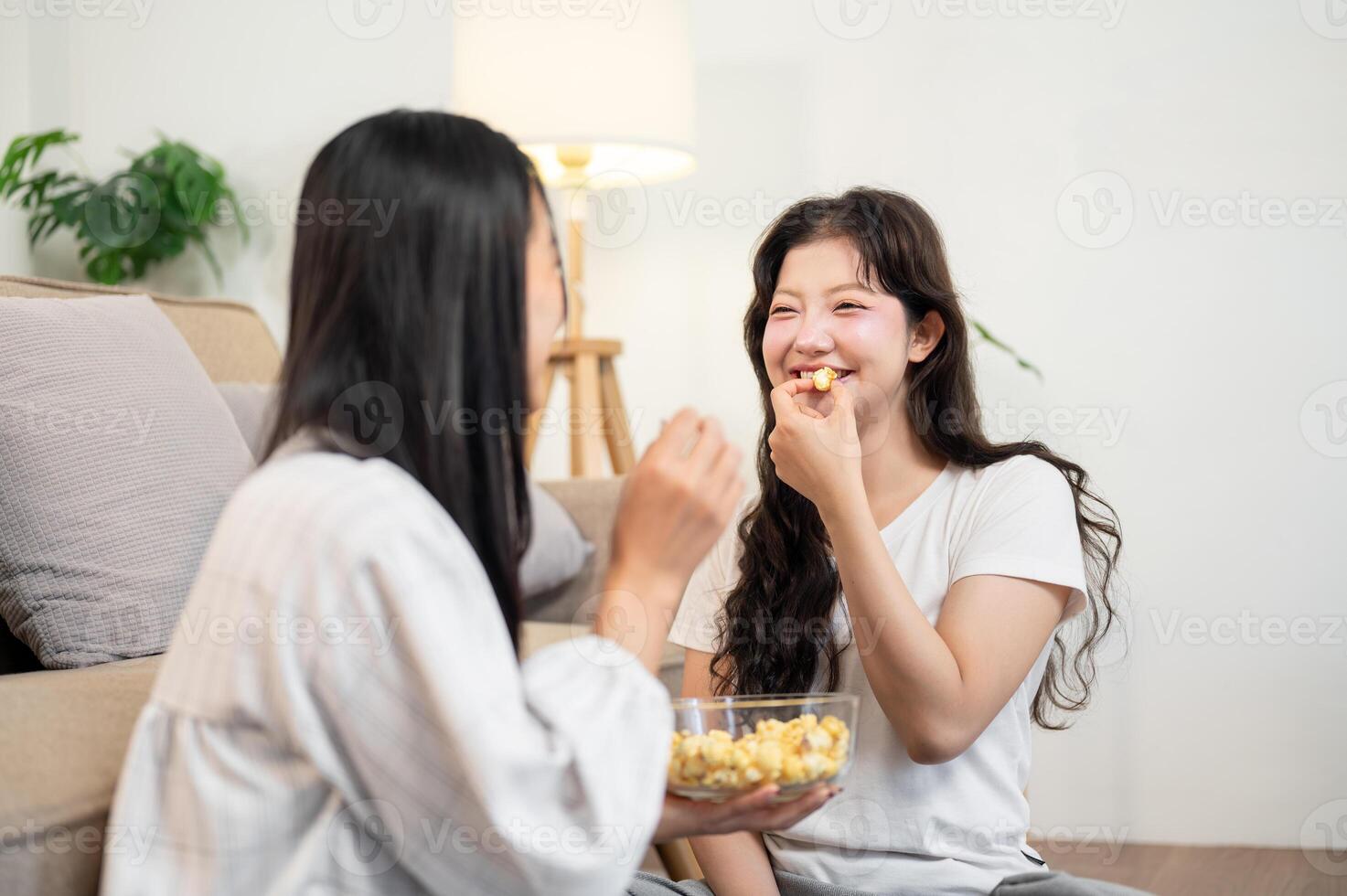 Two happy young Asian women are enjoying popcorn and a conversation together in the living room. photo