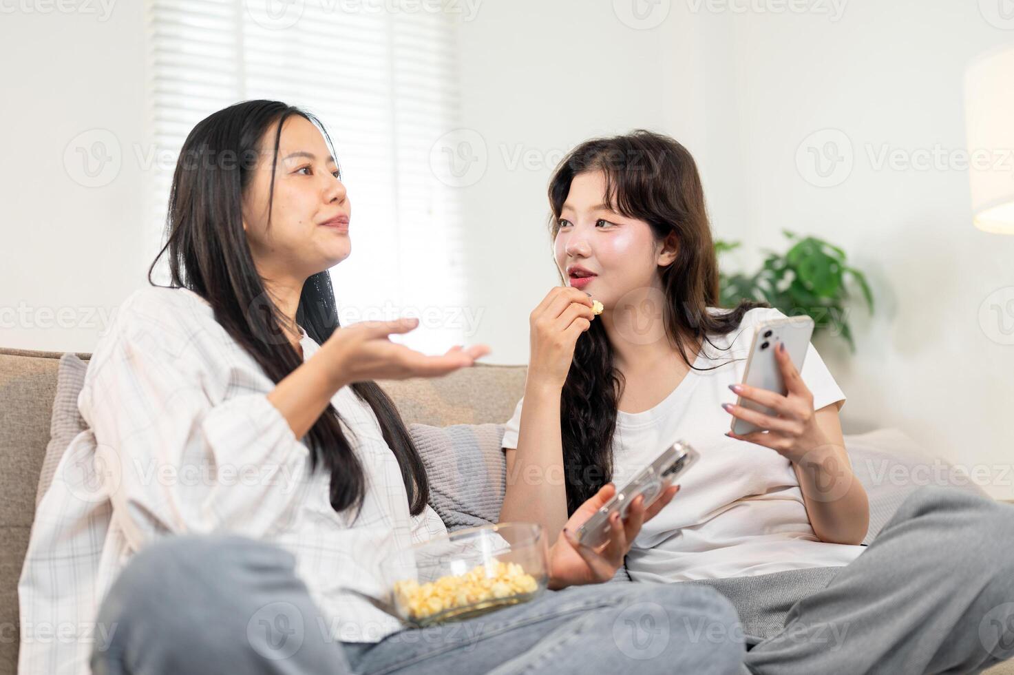 Two happy Asian female friends sit on a sofa, enjoying snacks and sharing their stories. photo