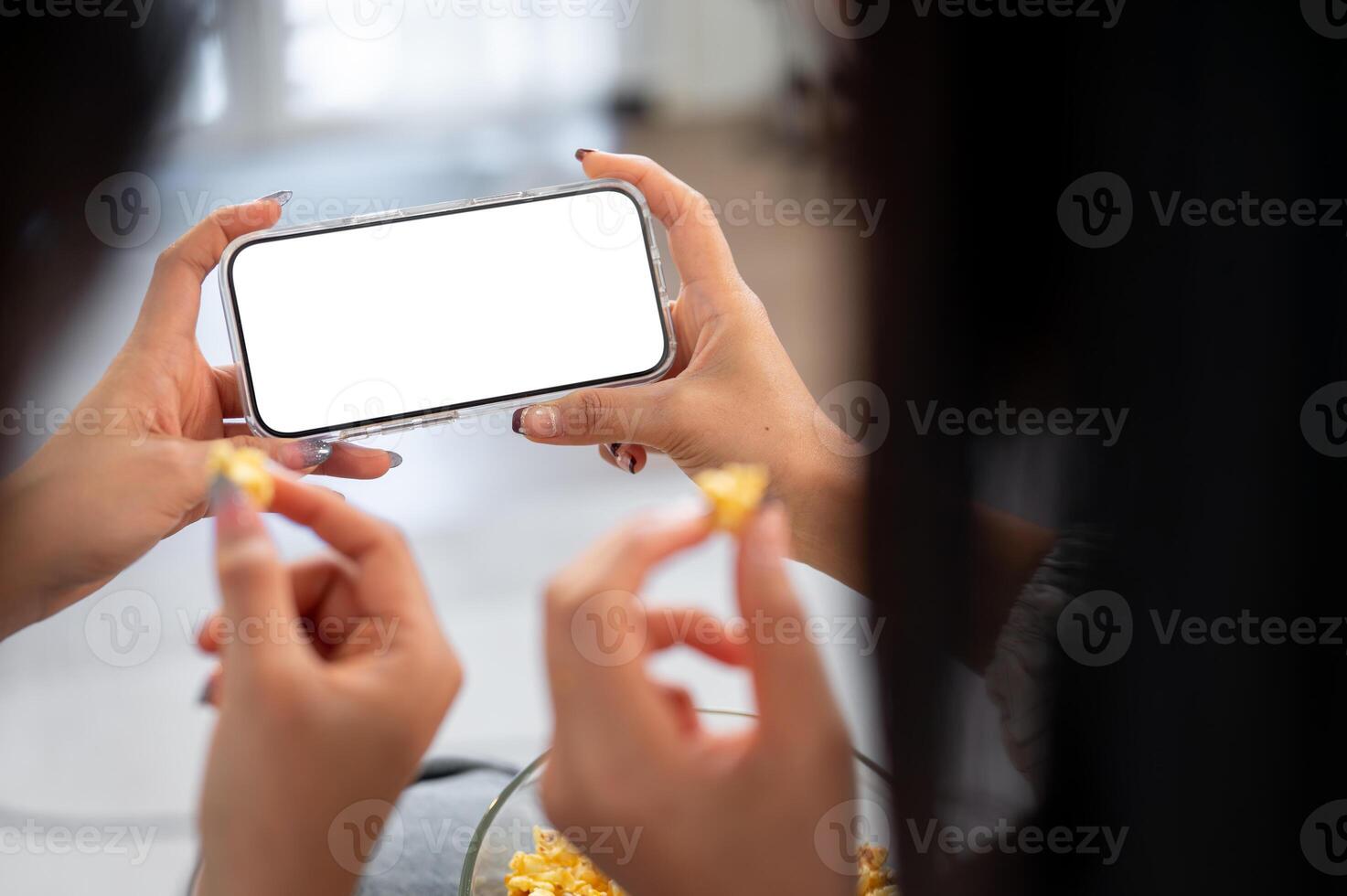 Two female friends chatting and snacking while watching a movie on a phone together on a sofa. photo