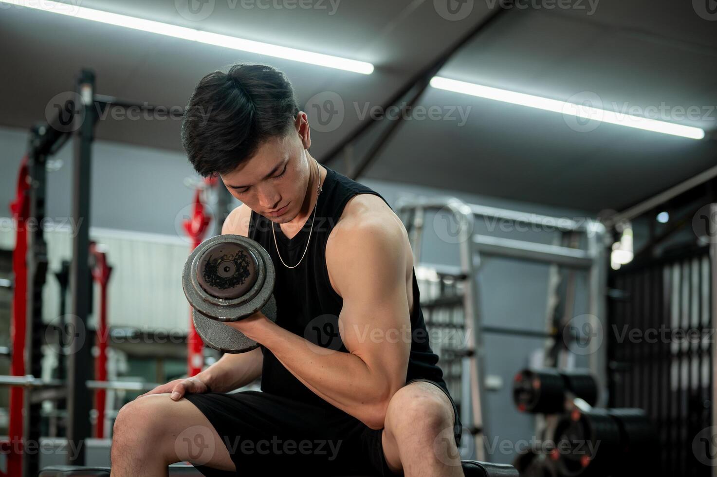 A muscular and strong Asian man in sportswear sits on a fitness bench, focused on lifting a dumbbell photo