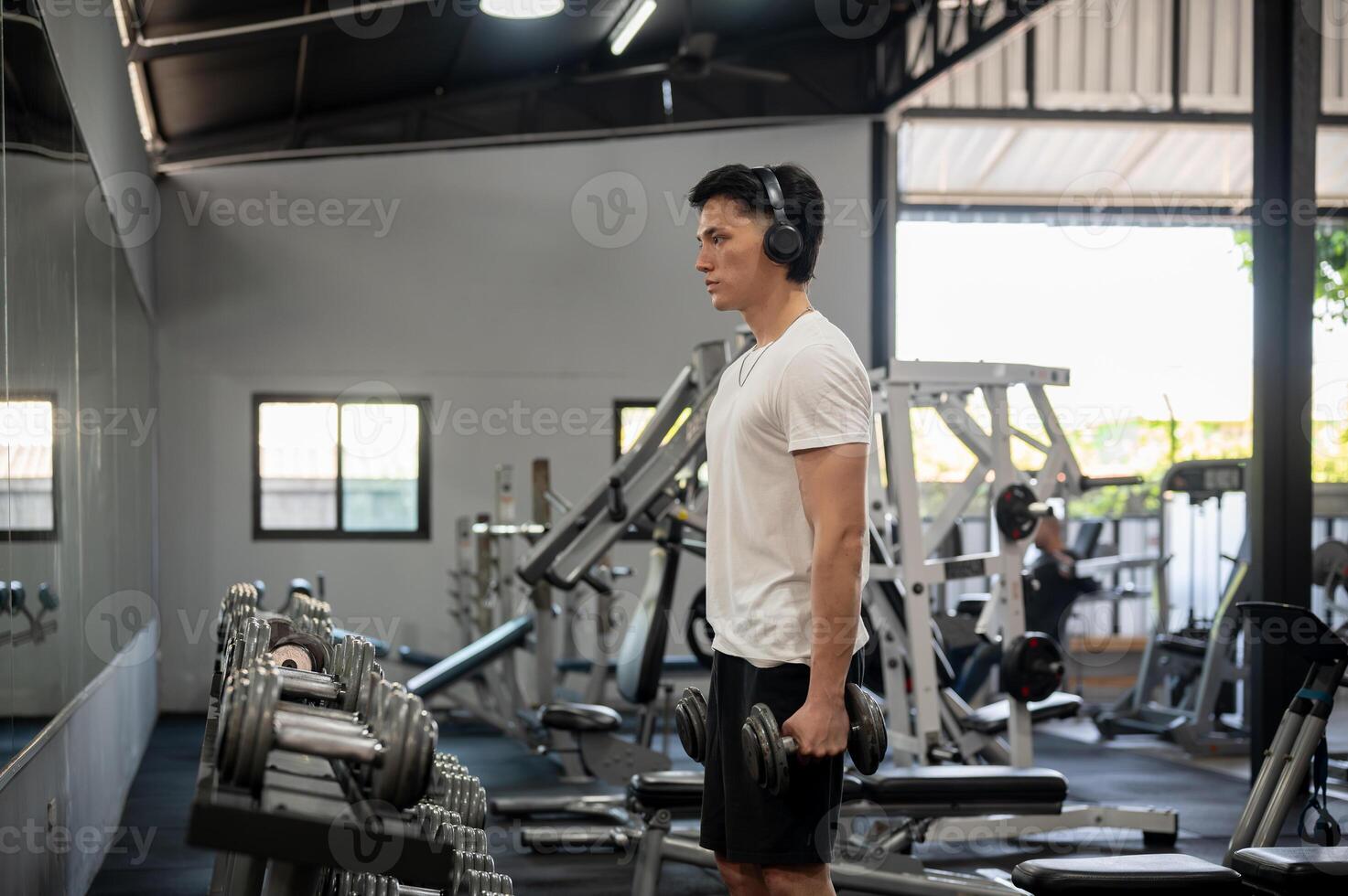 Focused Asian man is working out with dumbbells in front of the dumbbell rack and mirror in the gym. photo