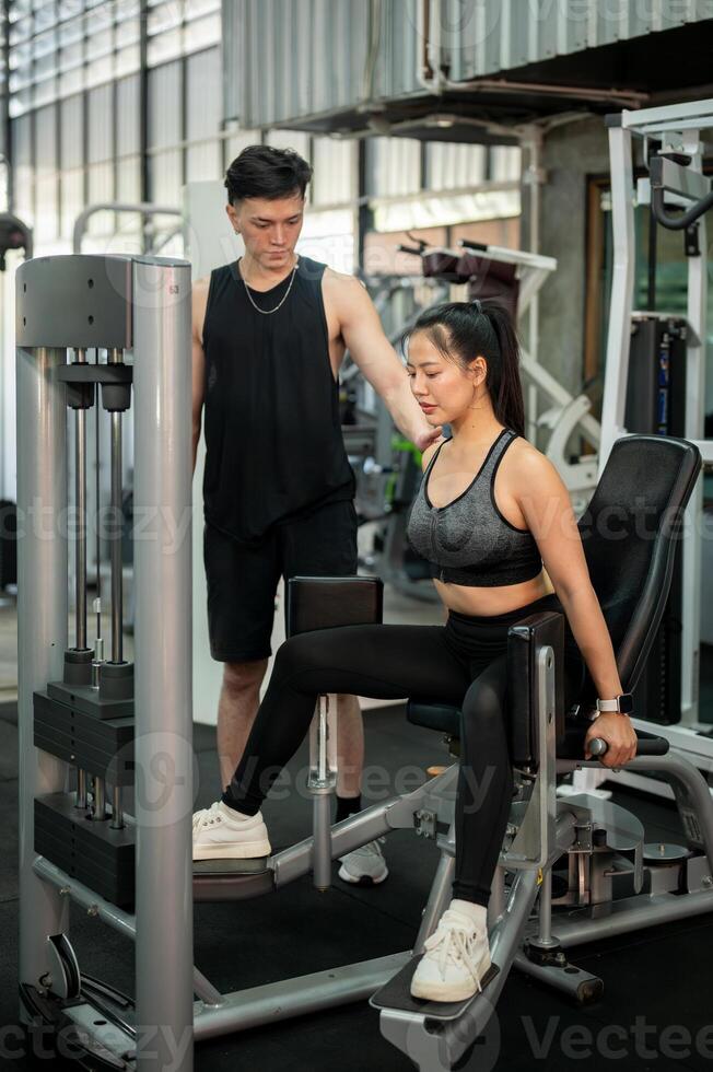 A focused Asian woman is being trained by a fitness coach, using the adductor machine in the gym. photo