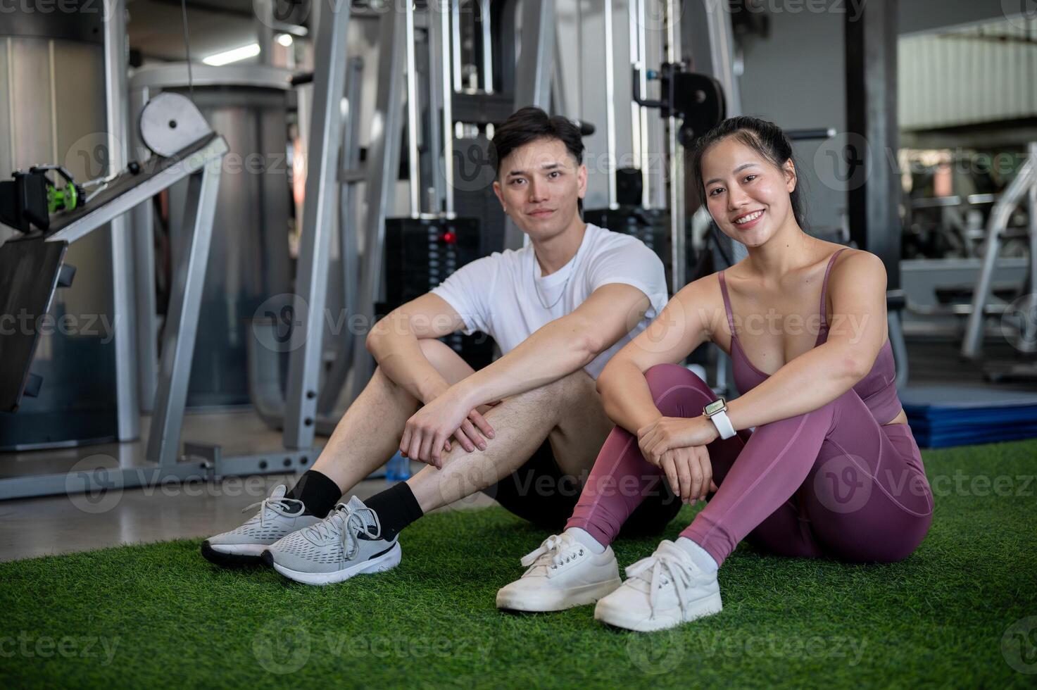 Sporty Asian male and female in sportswear are sitting on the floor of a gym, smiling at the camera. photo