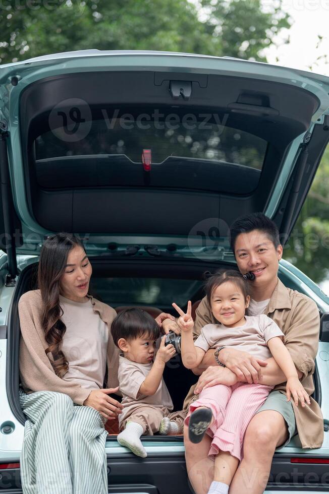A cheerful Asian family sitting in the trunk of their car, smiling and enjoying their road trip. photo