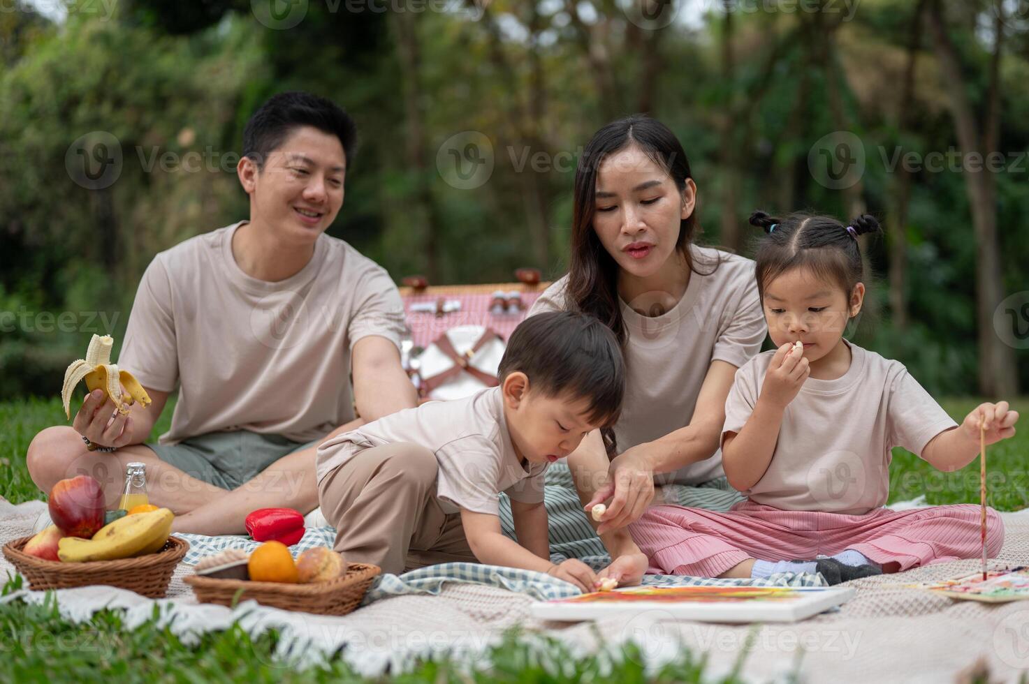 A cheerful Asian family enjoys a picnic in a green public park, sitting on a blanket, and eating. photo