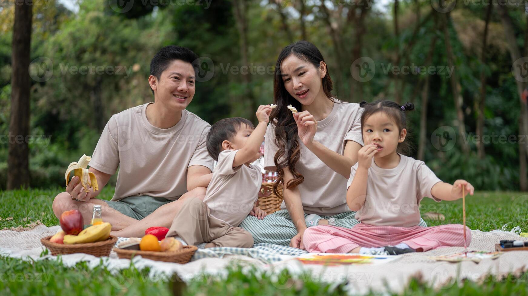 A happy Asian family enjoys a picnic in a green public park, sitting on a blanket, and eating. photo