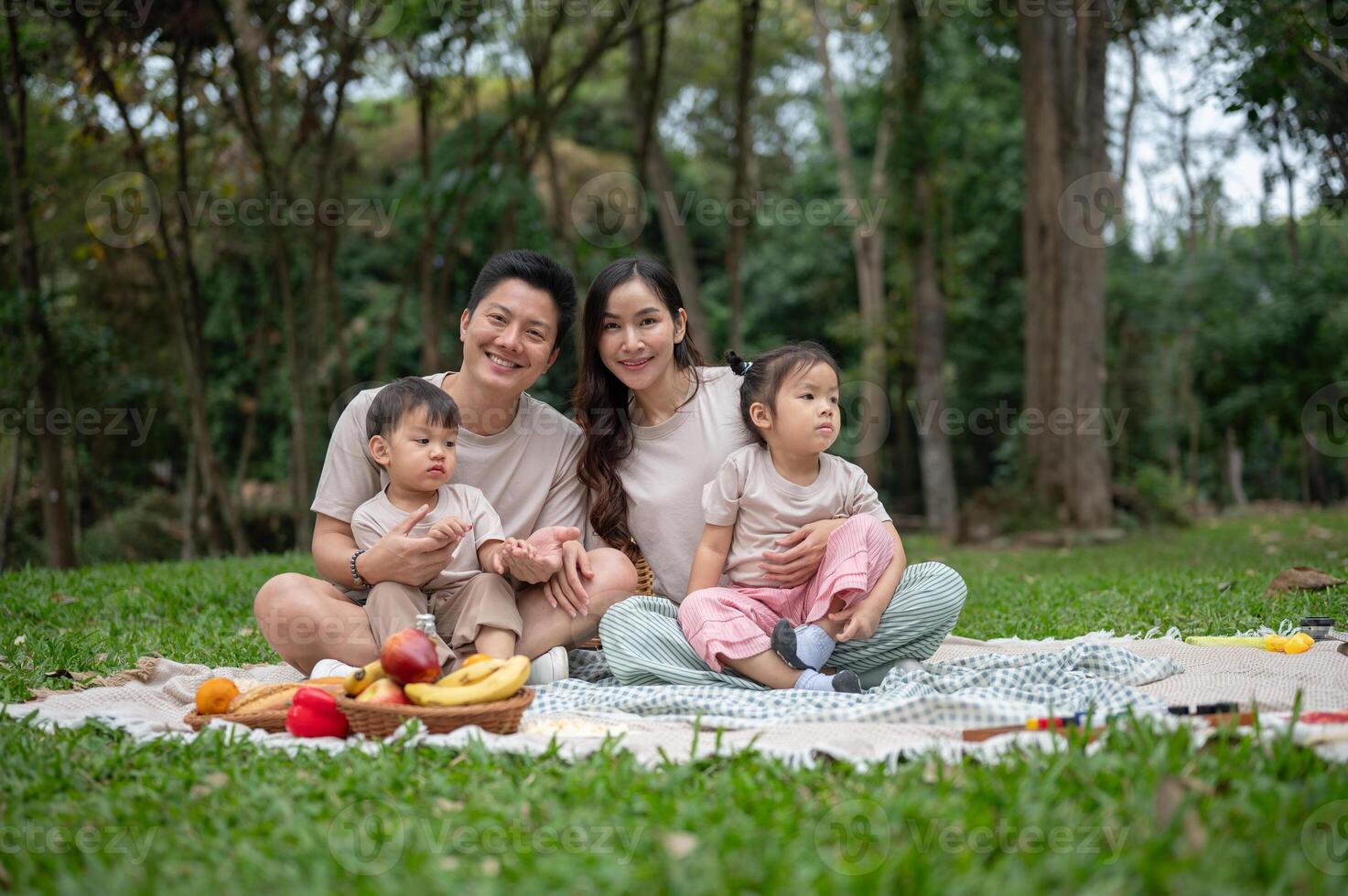 A happy Asian family sits on a picnic blanket in a green park, smiling at the camera. photo