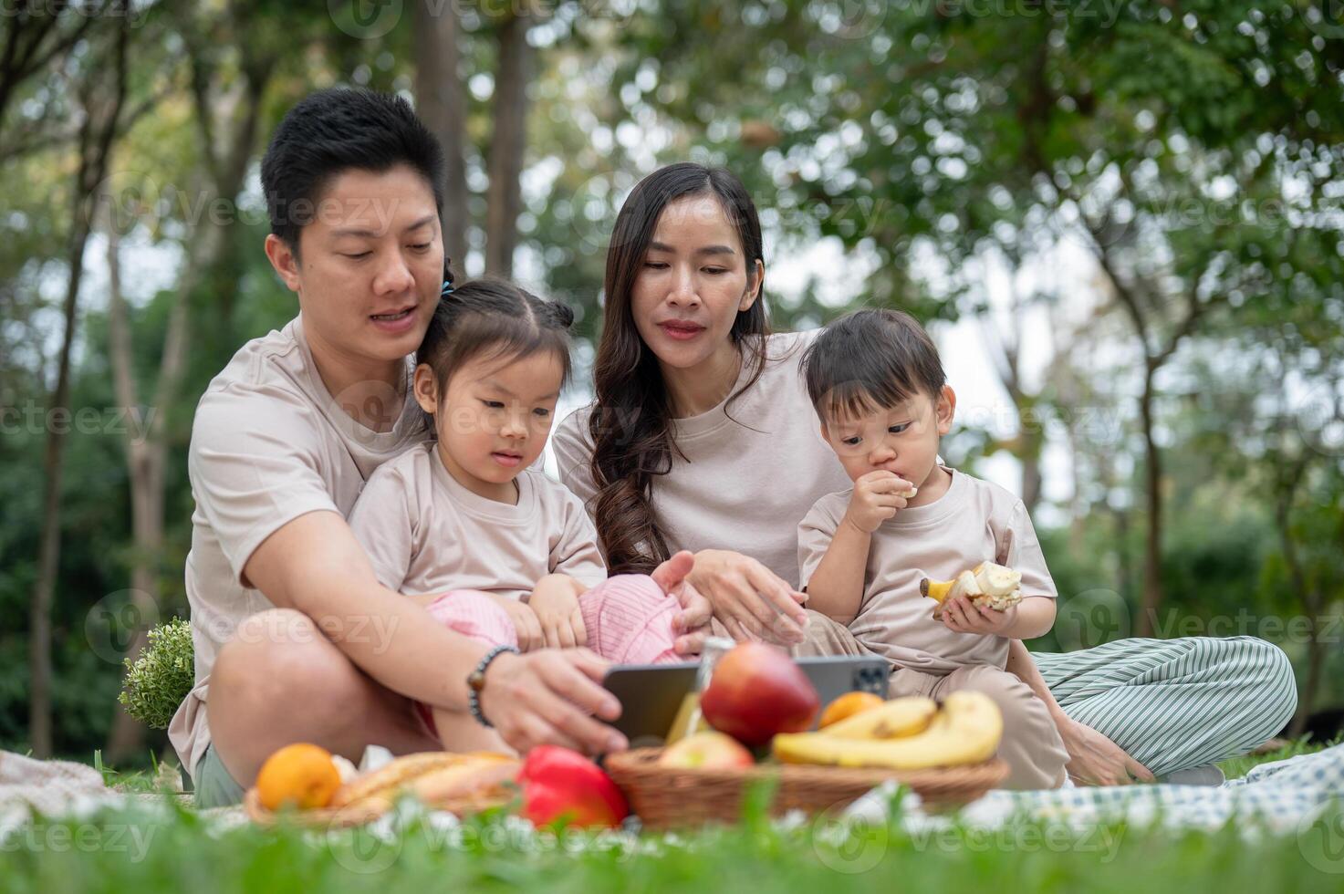 A happy Asian parents are taking their kids on a picnic in a park, watching a cartoon on a tablet. photo