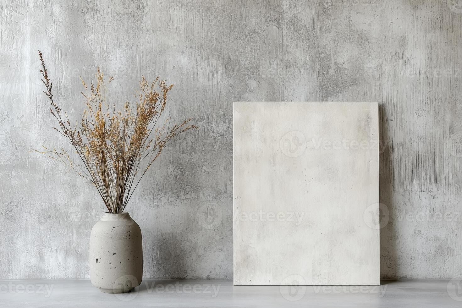 A neutral-toned vase with dried flowers placed next to a blank canvas, set against a textured gray wall, showcasing simple and elegant decor. photo