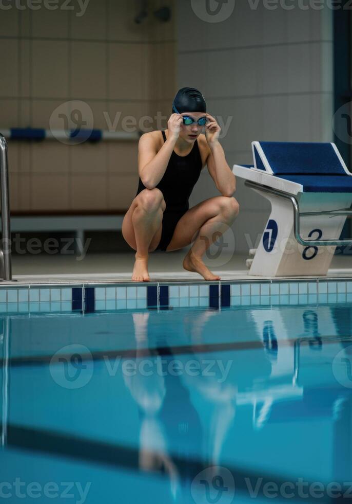 Female swimmer preparing to dive at indoor poolside in black swimsuit and goggles photo