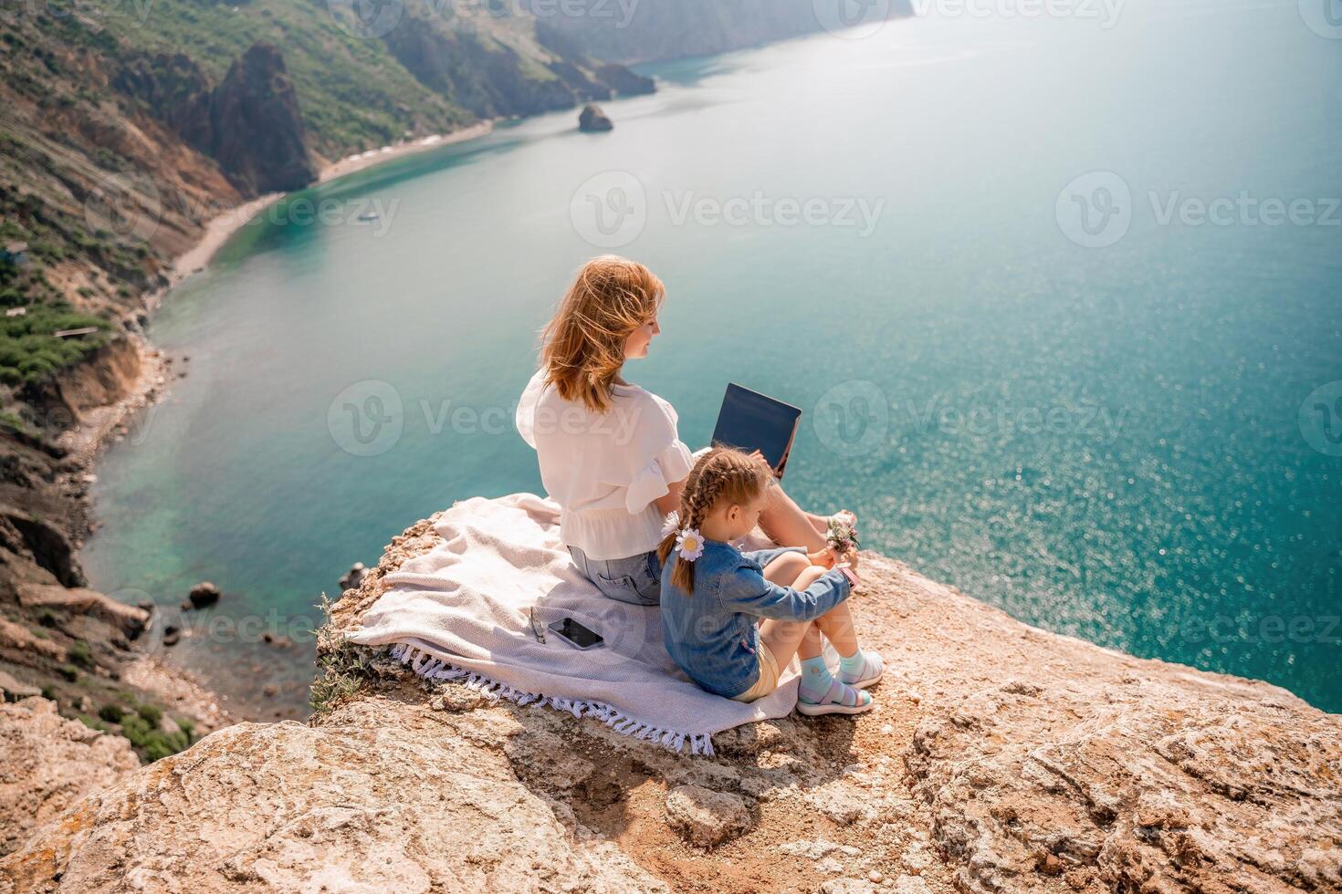 Freelance woman with her daughter working on a laptop by the sea, typing on the keyboard, enjoying the beautiful view, highlighting the idea of remote work. photo