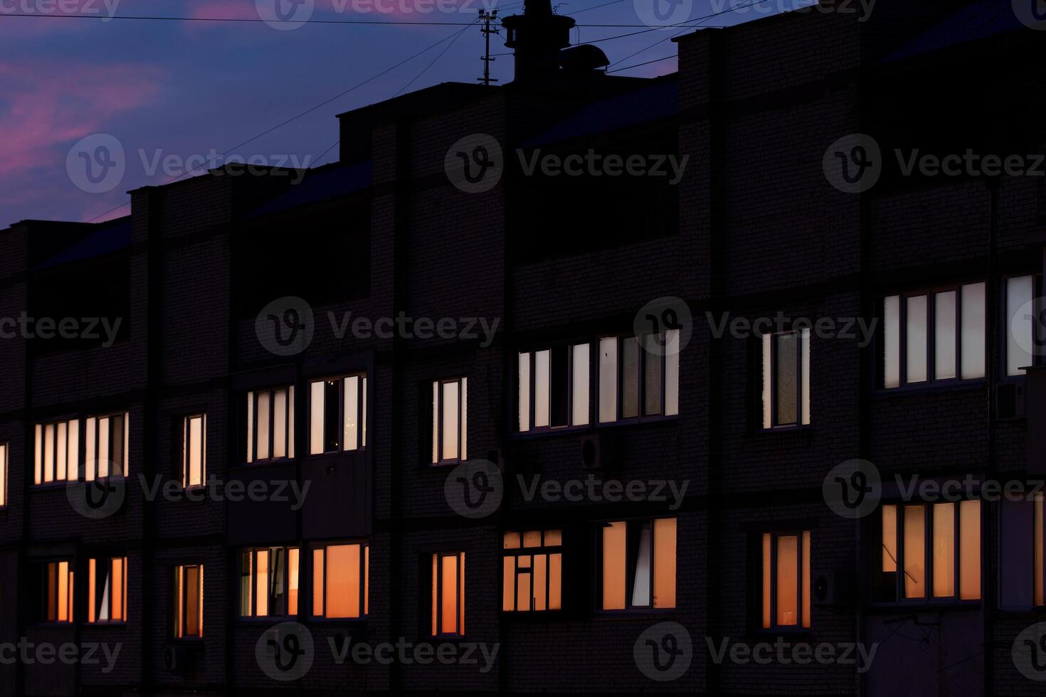 A multi-story brick building at dusk with illuminated and dark windows. Antennas and rooftop structures are visible. The sky transitions from deep blue to soft pink, creating a twilight ambiance. photo