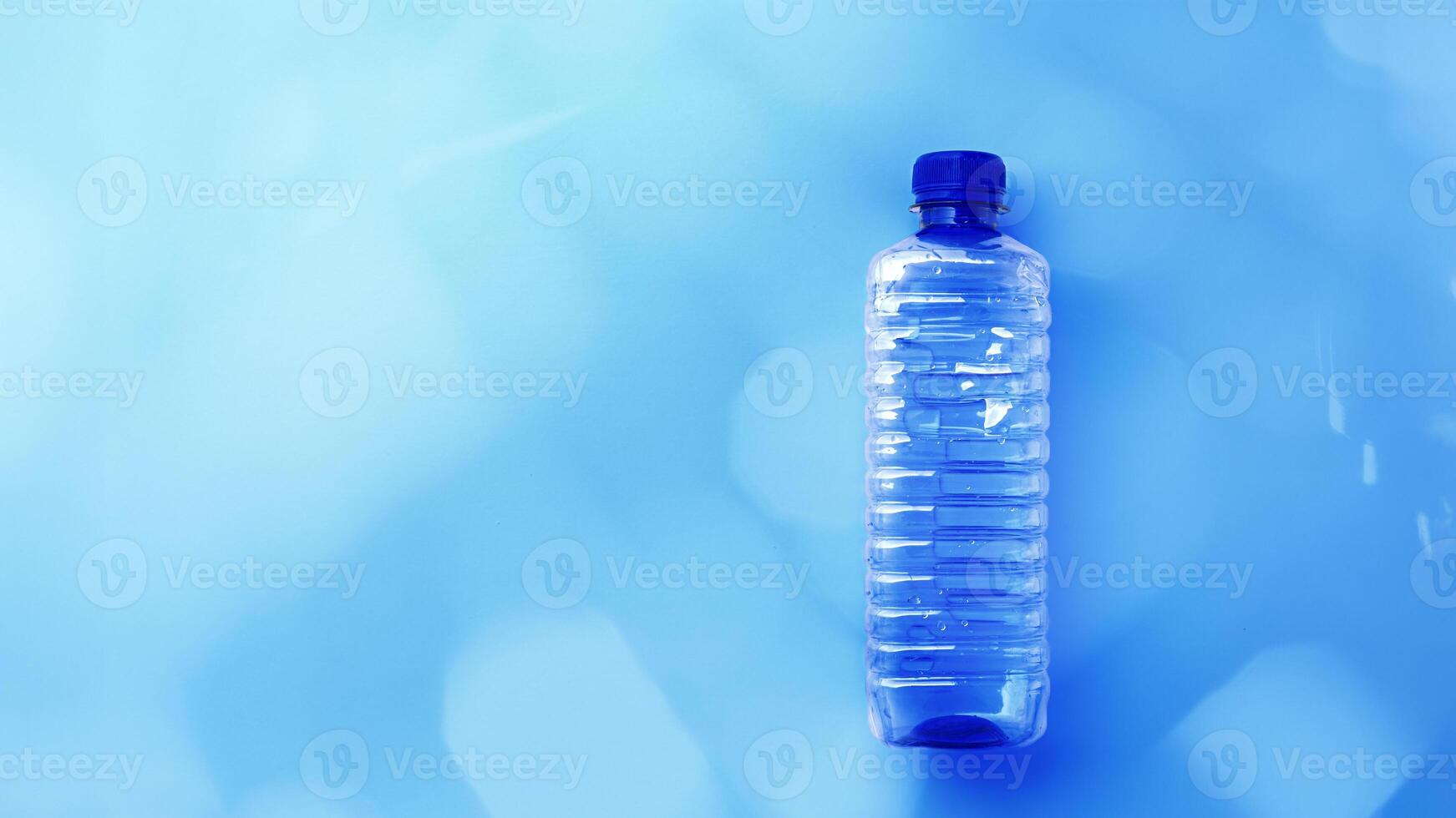 Clear plastic water bottle on bright blue background with light reflections, color gradient banner with copy space. Minimal style top view photo, one transparent blue bottle, overview flat lay photo