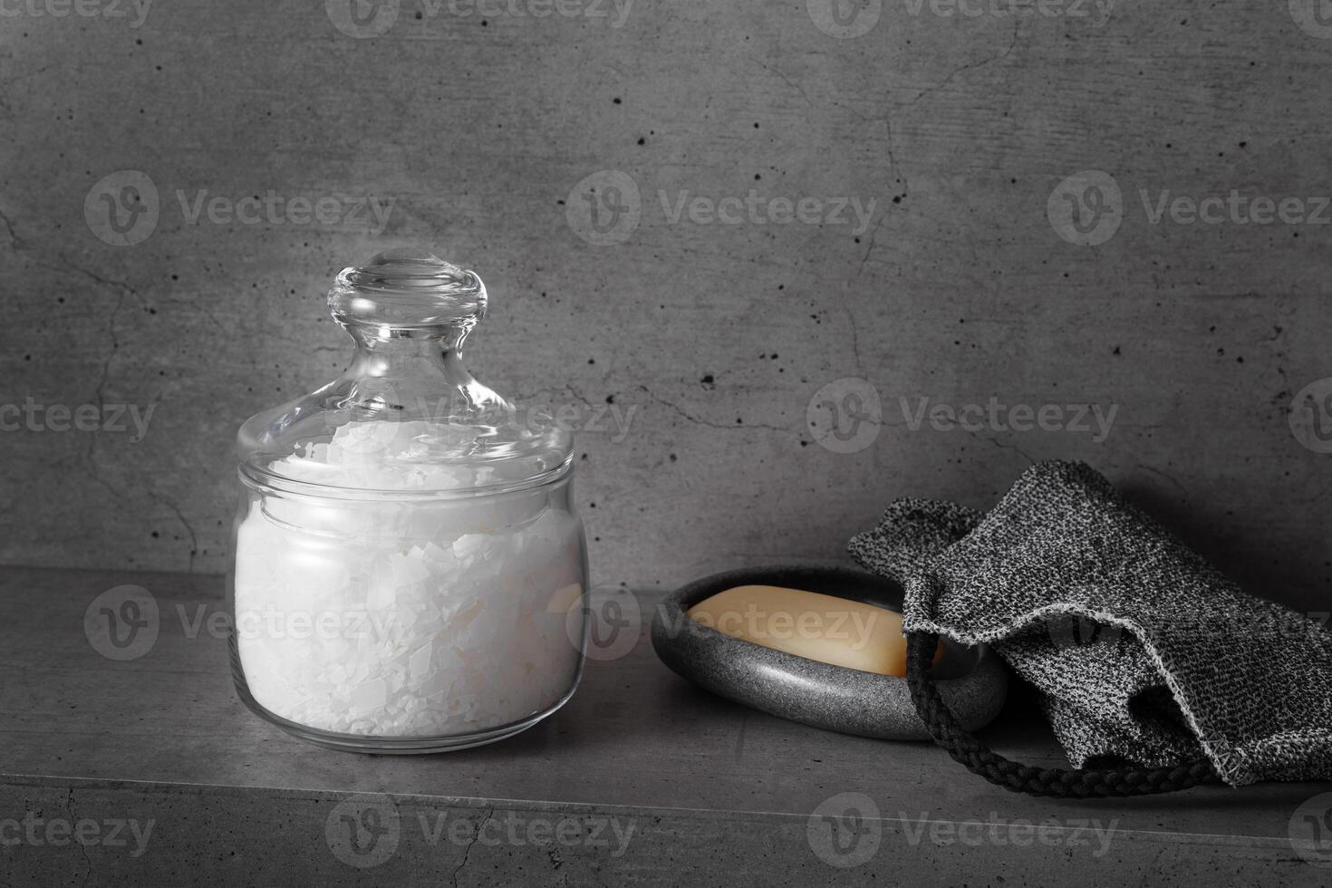 Glass jar with natural sea salt in grey bathroom, stone soap dish with soap, exfoliating body washcloth. Modern eco-friendly approach to self-care and wellness routines, home spa routine photo