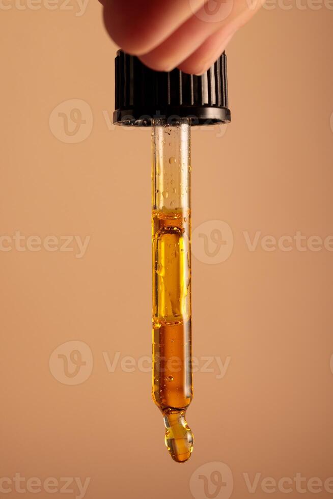 The hand is holding a pipette from which drops of yellow oil are dripping photo