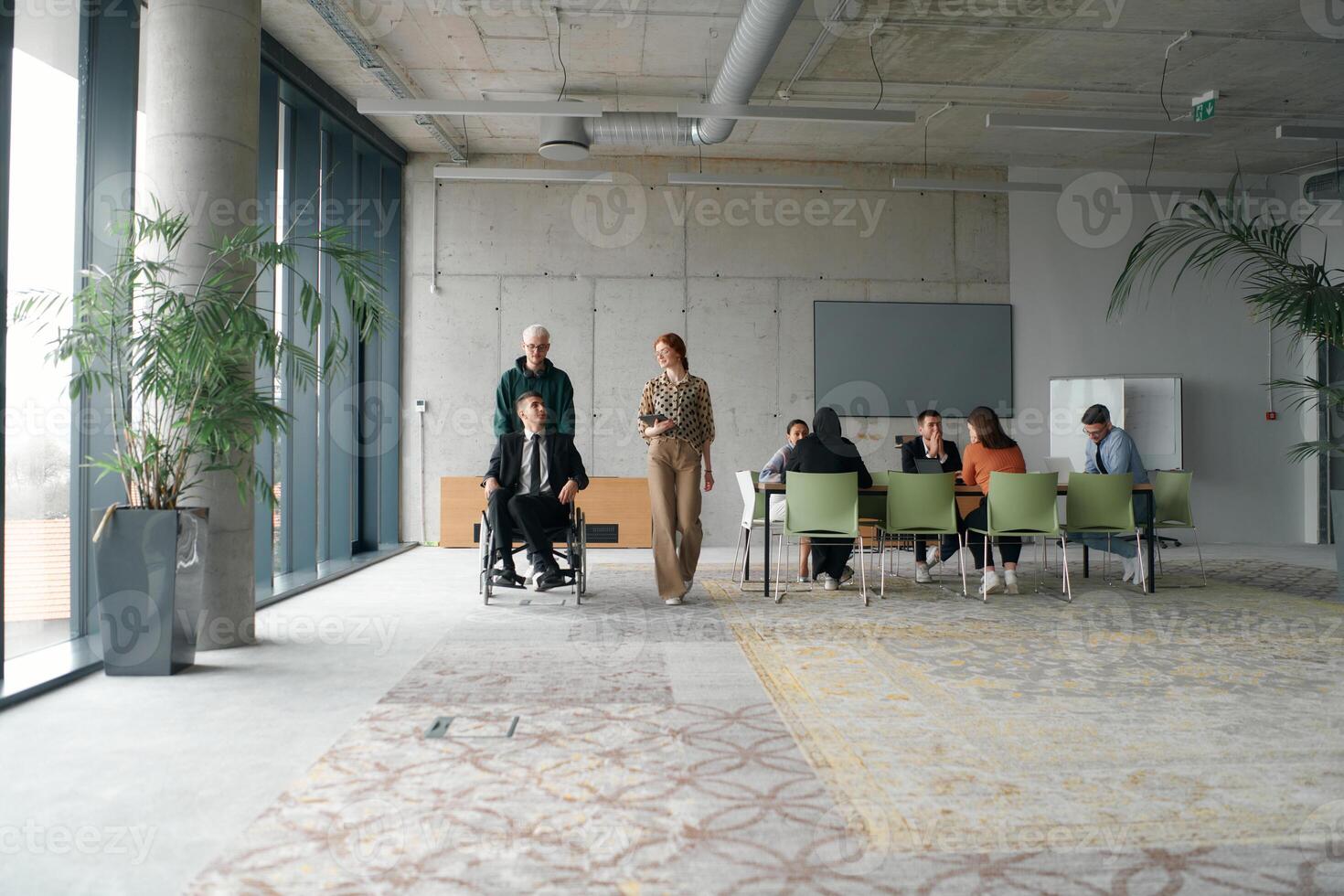 A diverse and multicultural business team collaborates in the office while their colleagues, including a director in a wheelchair and his friend, share a lighthearted moment, fostering an atmosphere photo