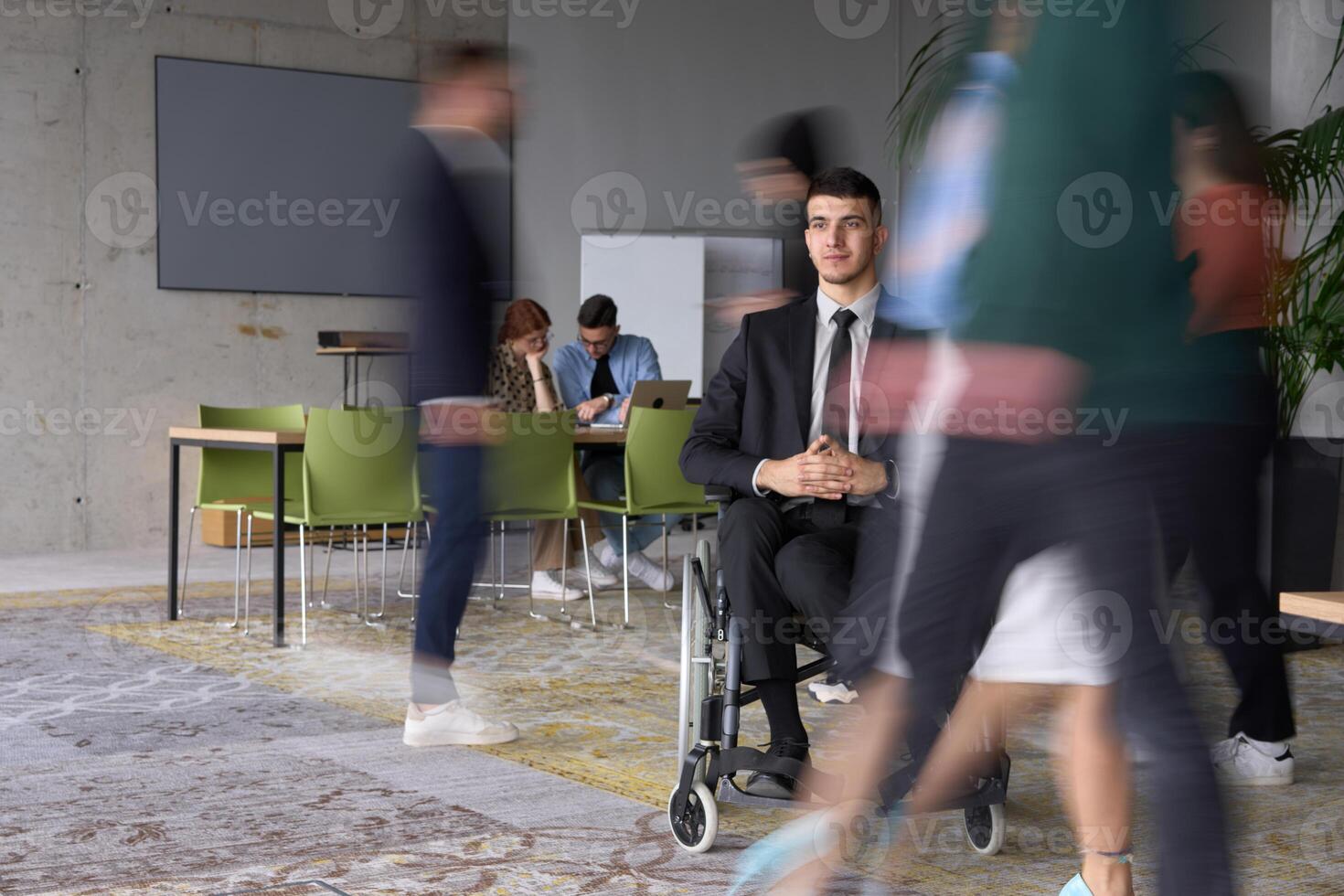 A businessman in a wheelchair navigating through a busy office, surrounded by his colleagues who are actively engaged in their work and collaboration photo