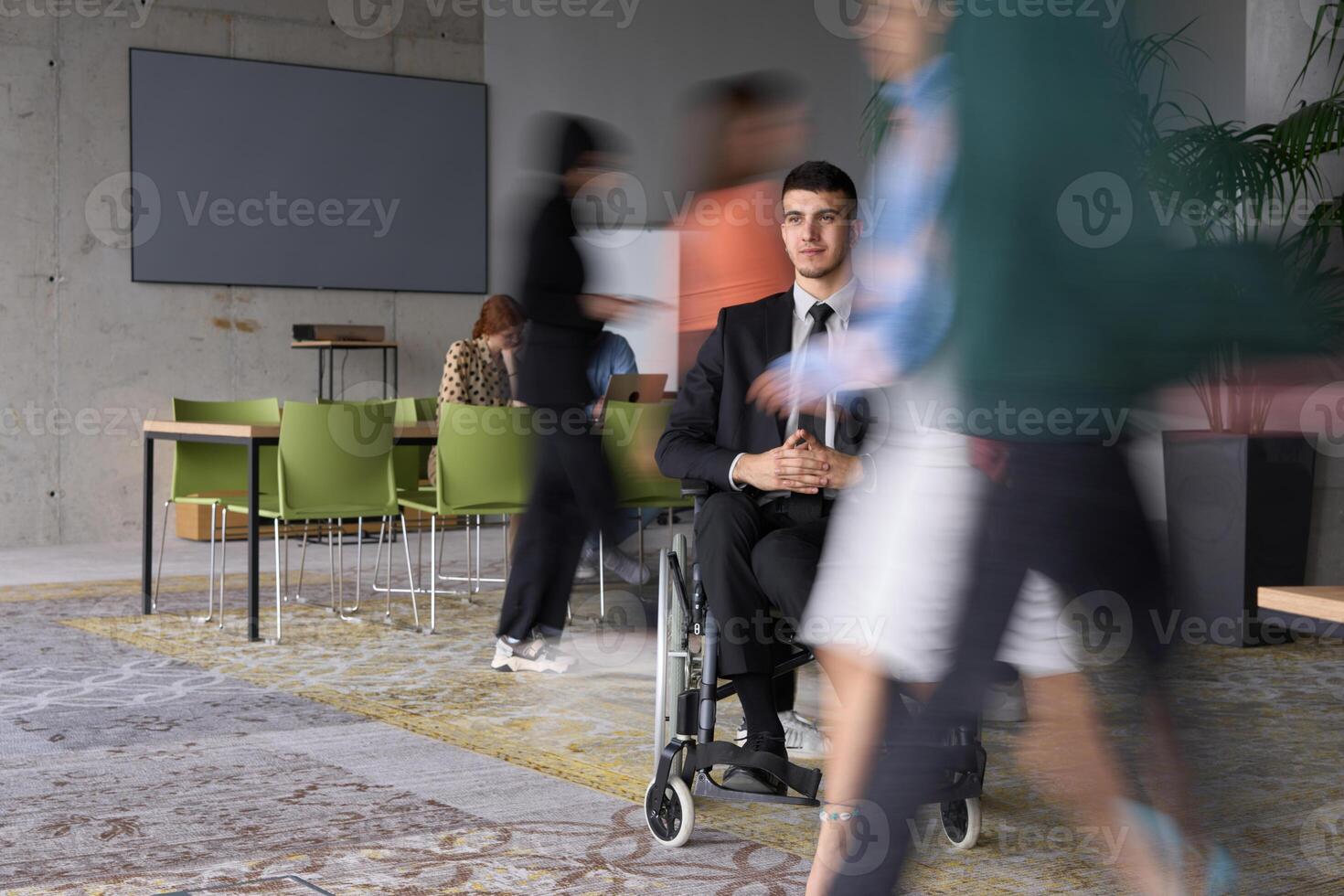 A businessman in a wheelchair navigating through a busy office, surrounded by his colleagues who are actively engaged in their work and collaboration photo