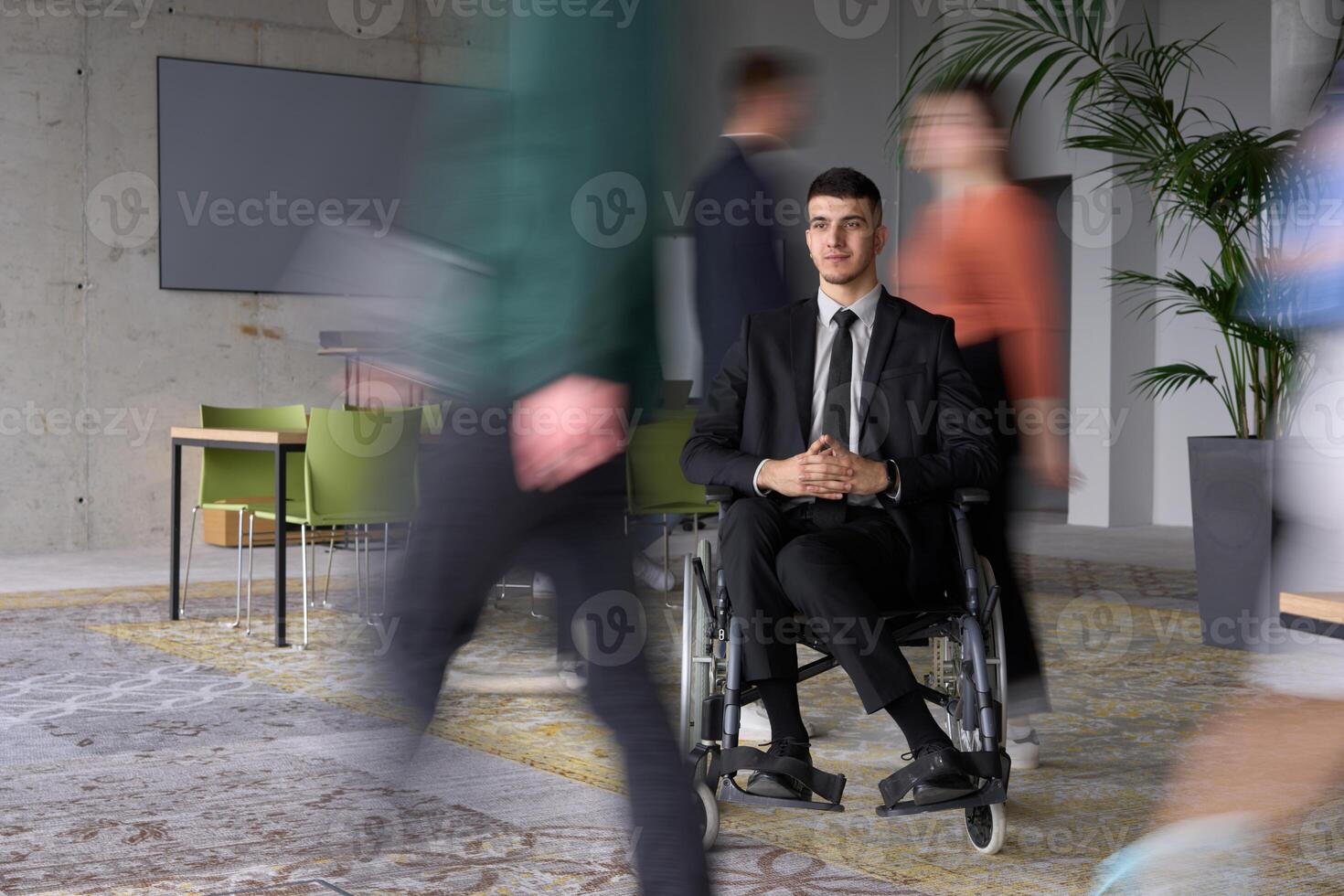 A businessman in a wheelchair navigating through a busy office, surrounded by his colleagues who are actively engaged in their work and collaboration photo