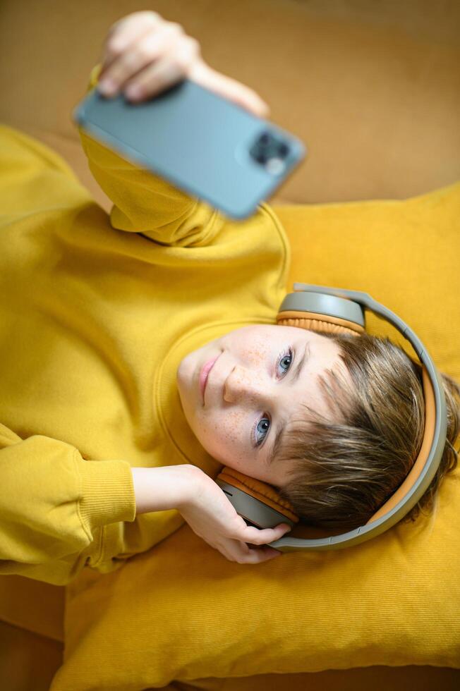 A boy lounges on a yellow sofa, headphones on, immersed in music on his smartphone photo