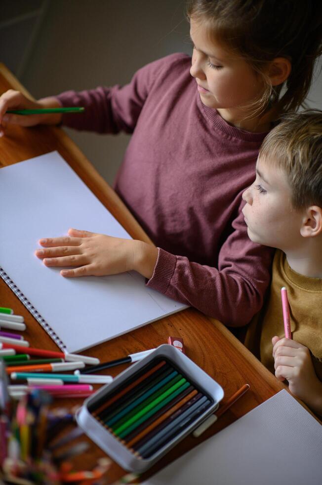 The siblings are surrounded by colorful art supplies, staring at their blank sheets of paper. photo