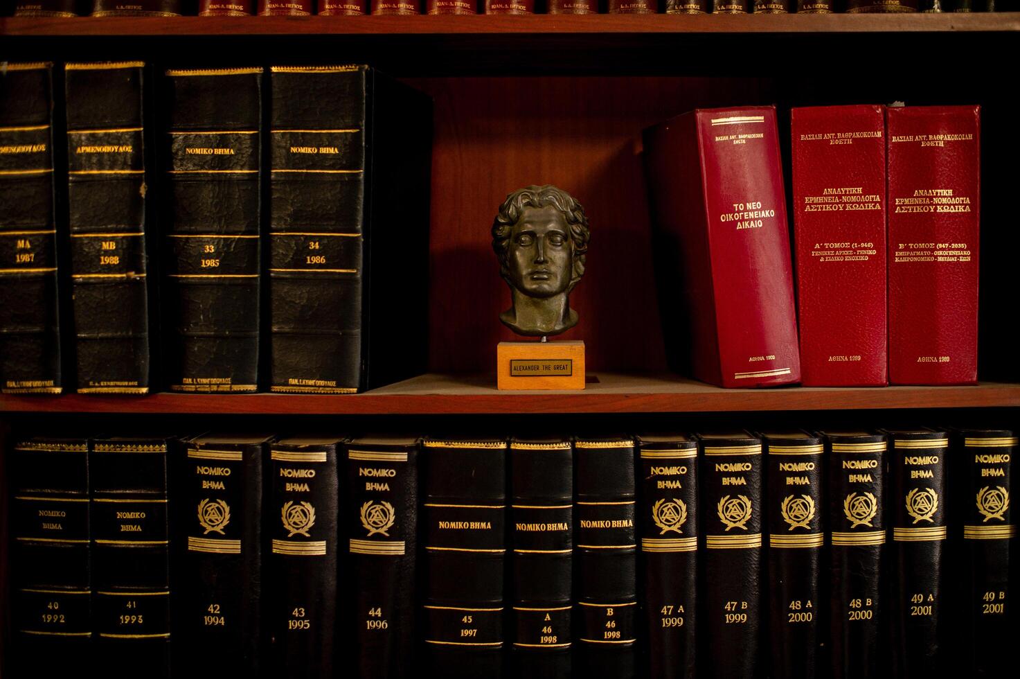 A wooden bookcase packed with legal books in Greek, with a marble bust of Alexander the Great prominently displayed. photo