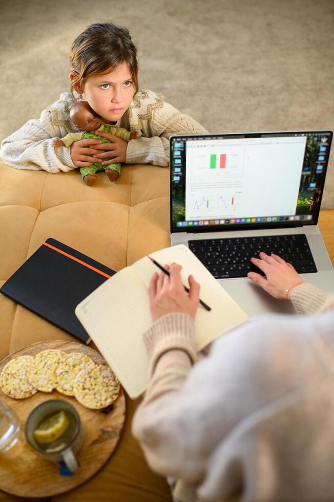 The mother is busy working, while the daughter waits with her doll, looking forward to playing together. photo