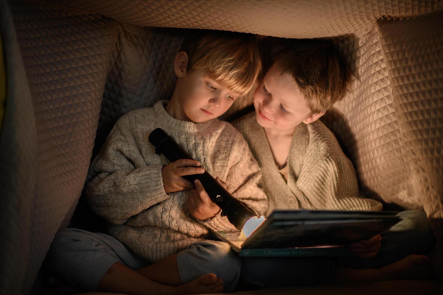 Two little boys reading a book under a blanket with a flashlight, creating a cozy, secretive atmosphere. photo