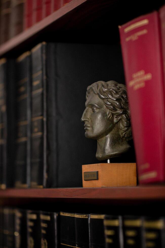 A wooden bookcase packed with legal books in Greek, with a marble bust of Alexander the Great prominently displayed.The combination of law and history creates a scholarly atmosphere. photo