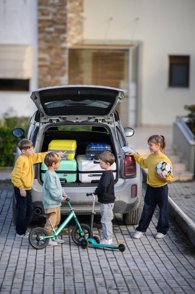 The open trunk of a car is surrounded by four kids organizing vibrant suitcases and scooters for their adventure. photo