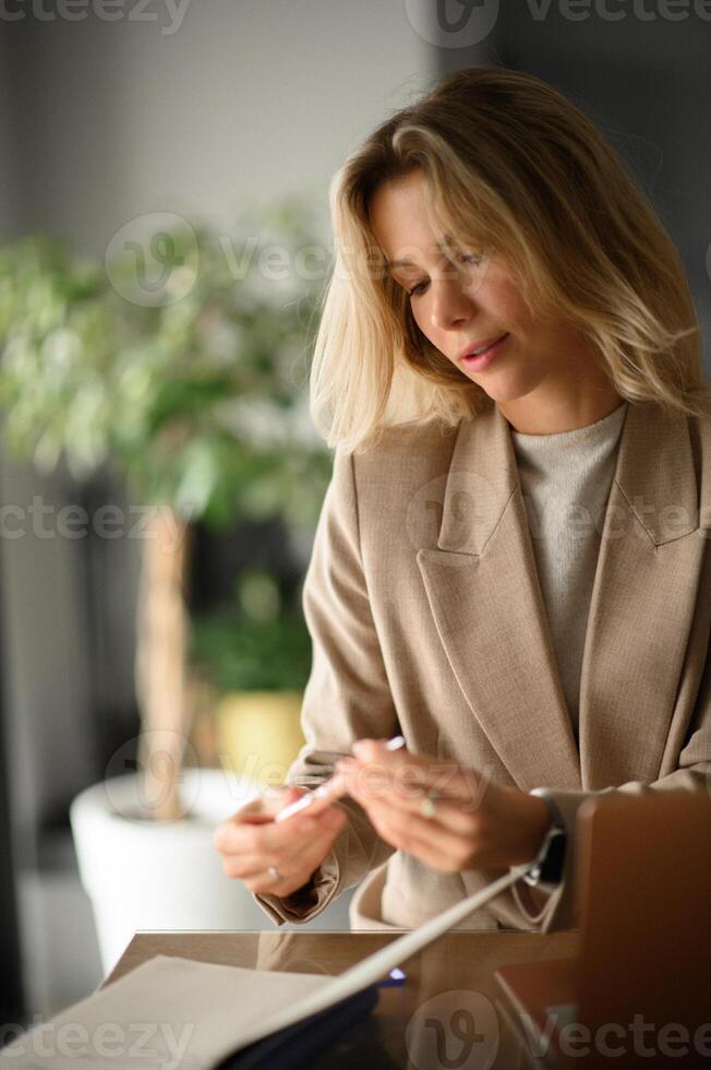 A blonde woman in a suit at an office desk, writing notes in a notebook with a focused expression. photo