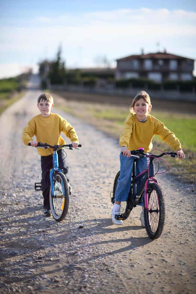 A girl and boy ride their bikes along a quiet rural road, surrounded by fields on a warm, sunny day. photo