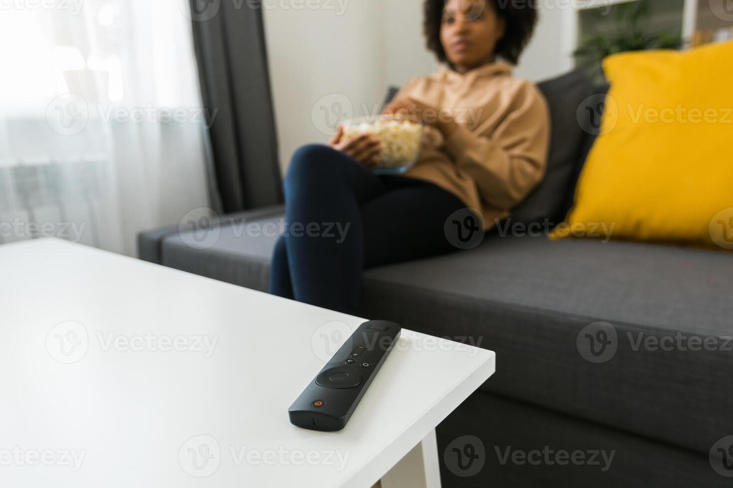 TV remote control on background of African american woman sits on a gray sofa, enjoying a bowl of popcorn while watching a movie or streaming online film. Cozy and relaxing moment of entertainment in photo