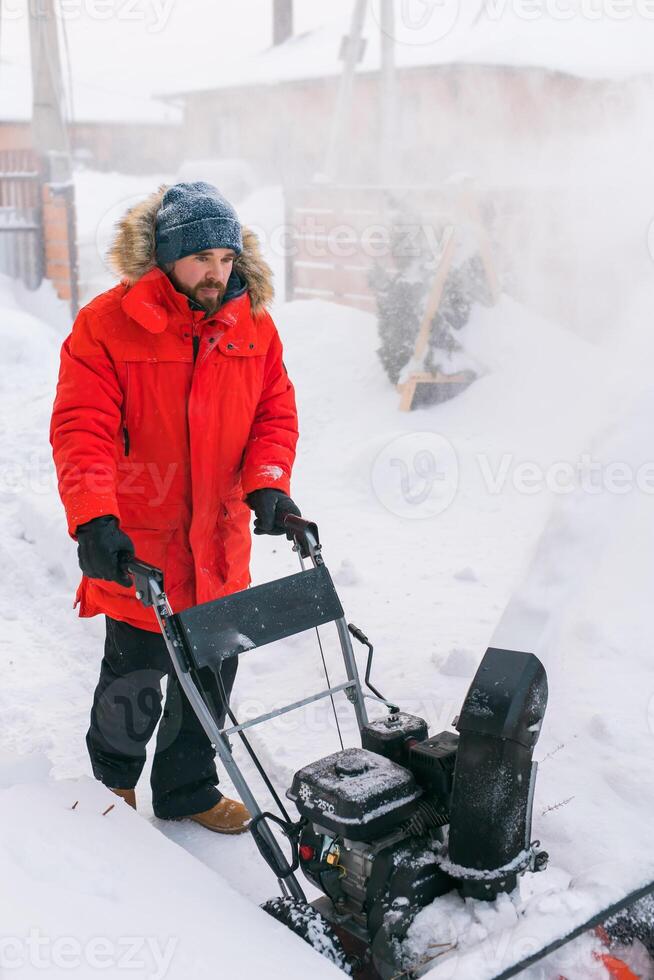Red snow blower in action. Man outdoor in front of house using snowblower machine for removing snow on yard. Snow thrower in winter outside home. Young worker guy blowing snow photo