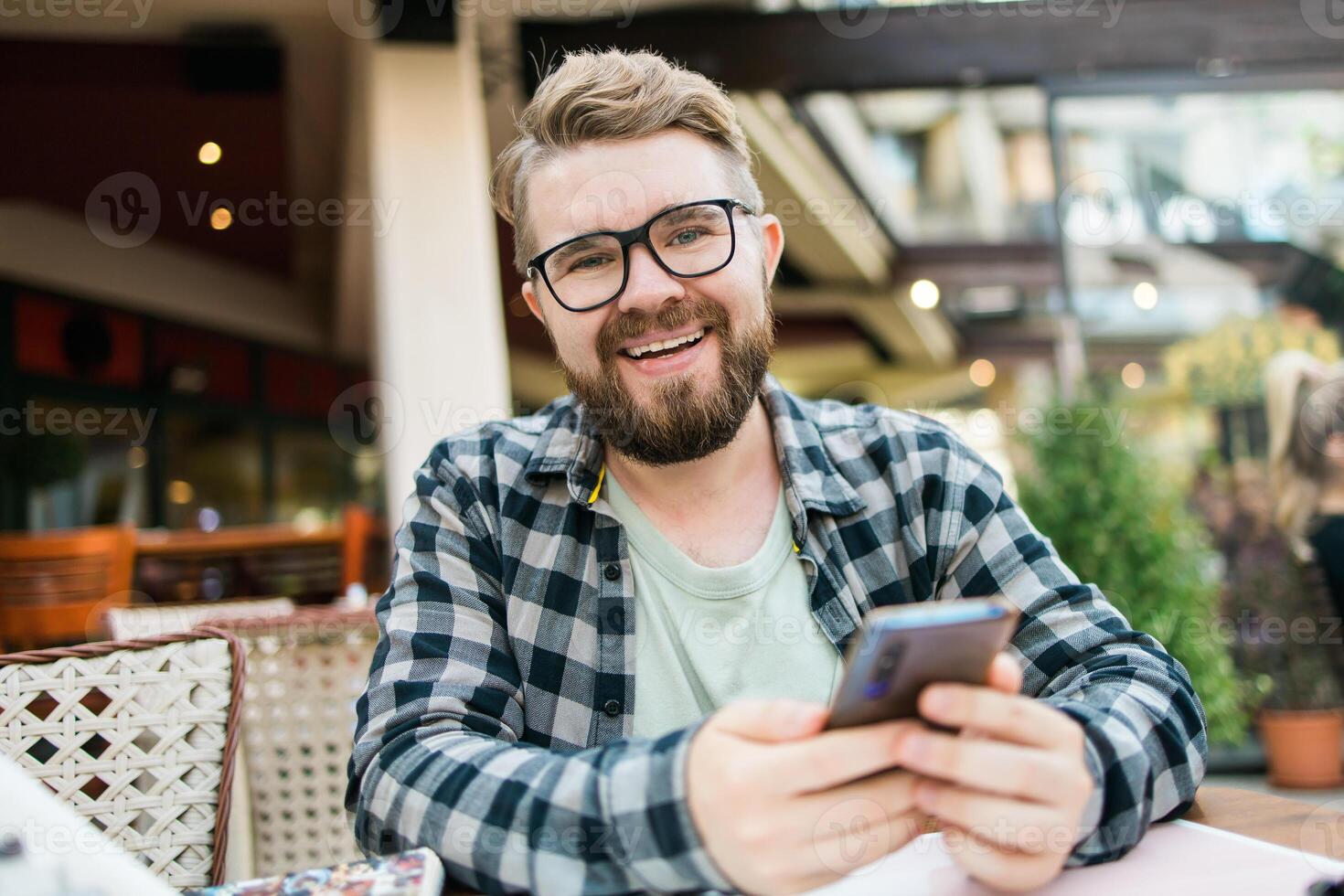 Smiling millennial man uses his smartphone while enjoying a casual outdoor cafe setting. Holidays atmosphere, modern connectivity, social networks photo
