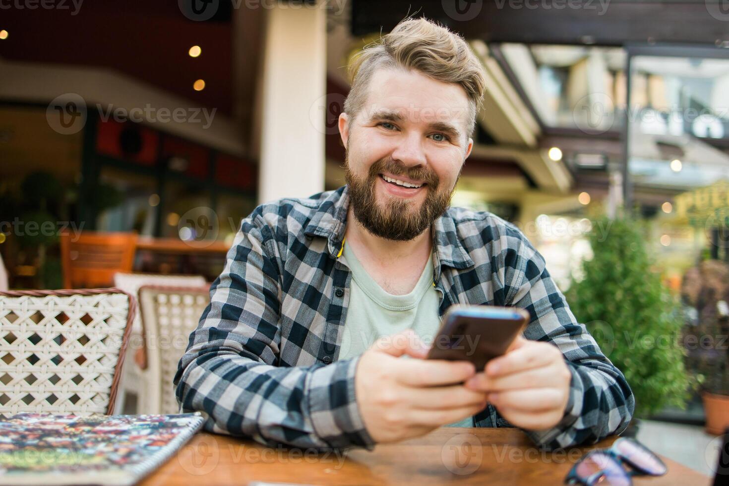 Smiling millennial man uses his smartphone while enjoying a casual outdoor cafe setting. Holidays atmosphere, modern connectivity, social networks photo