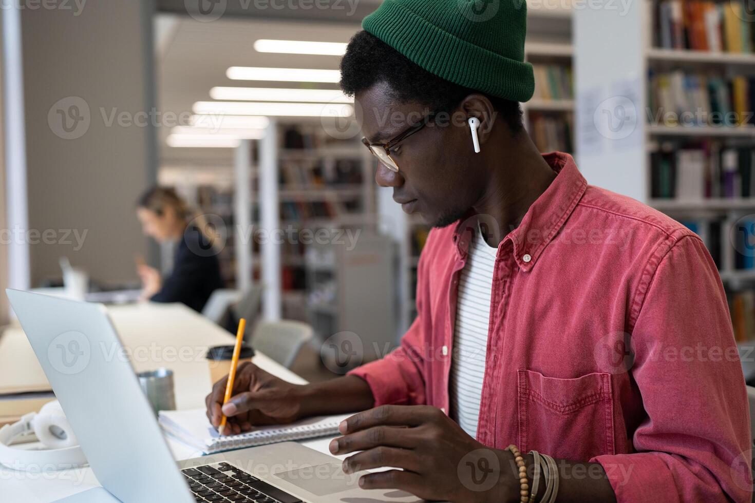 Focused emotionless African American man making notes in notebook sits at table with laptop photo