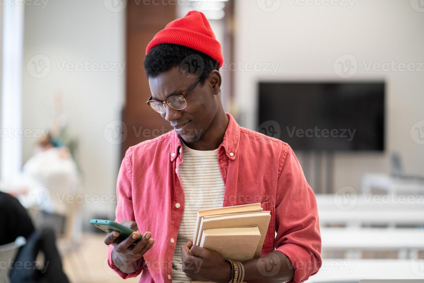 Satisfied hipster black student guy stands in college library with textbooks using phone break study photo