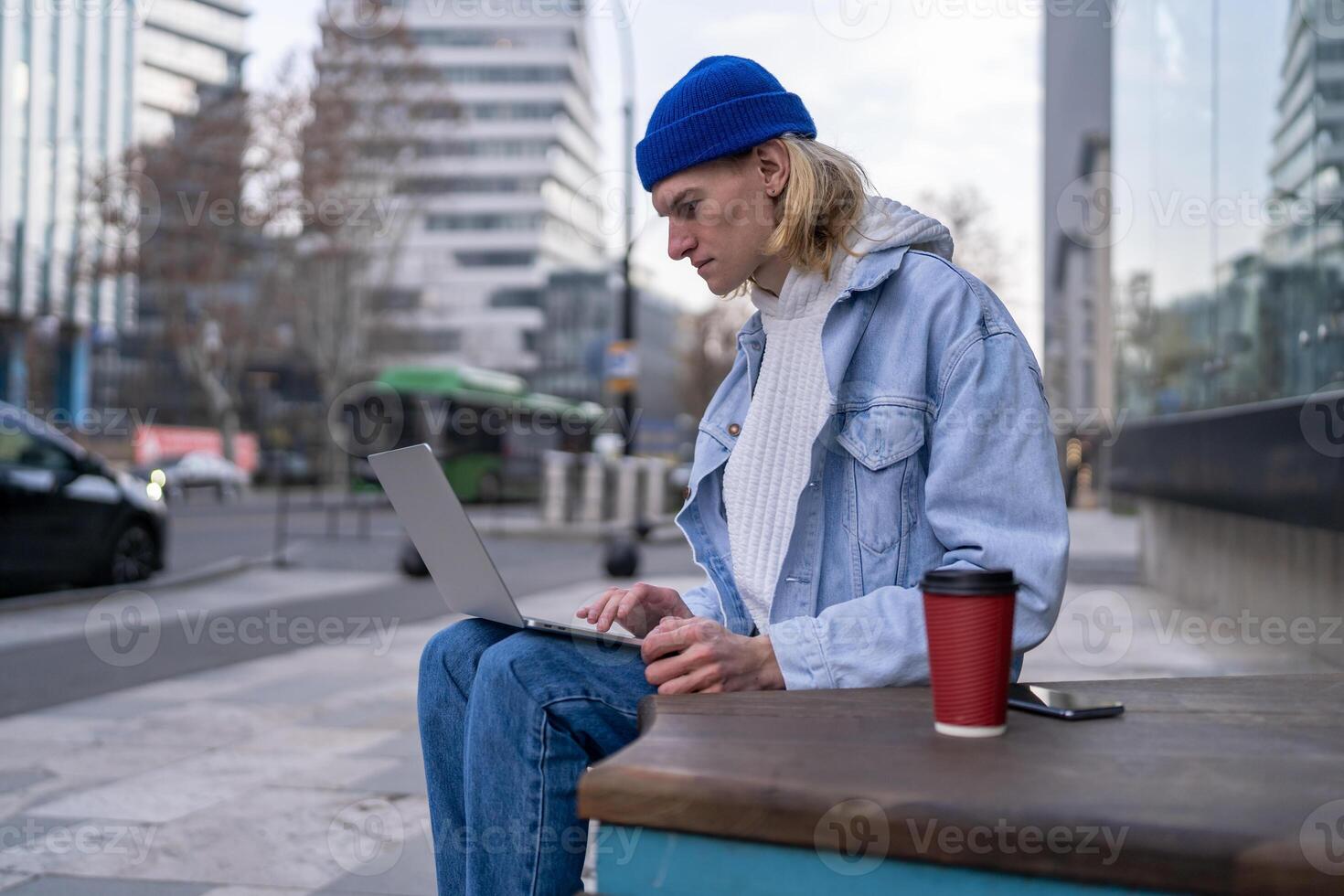 Fashionable guy sitting in urban street searching info in laptop computer, working, posting photos