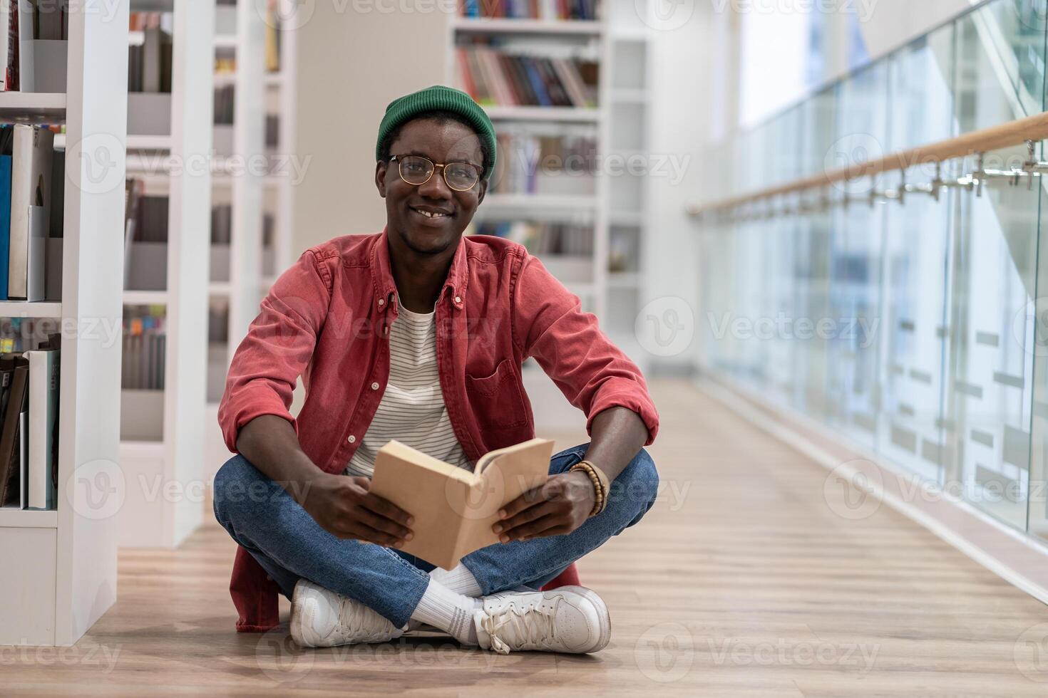 Happy clever black student man with book sitting on floor in university library, looking at camera photo