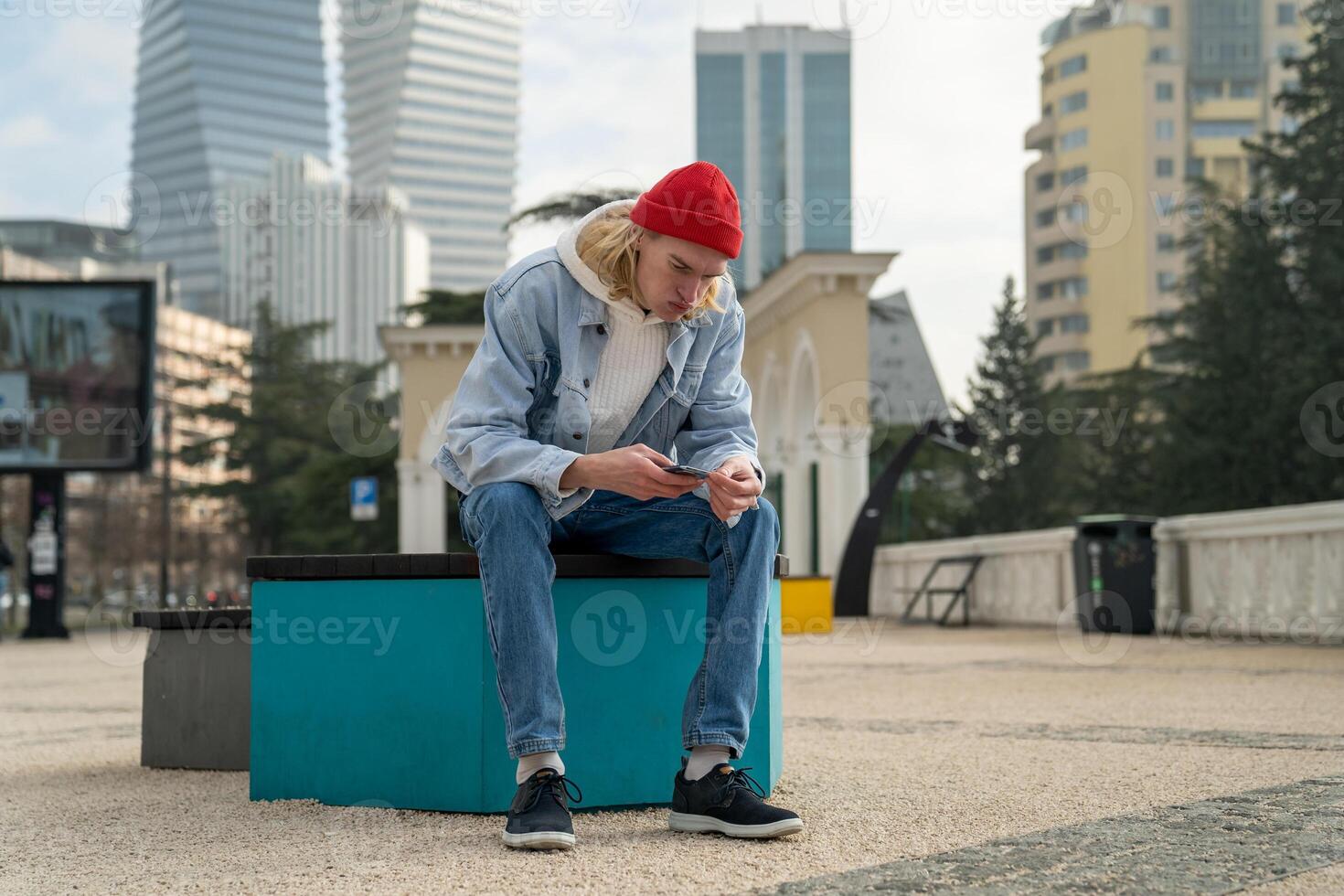 Pensive focused guy in red hat sitting outdoors, searching info in mobile phone, messaging chatting. photo
