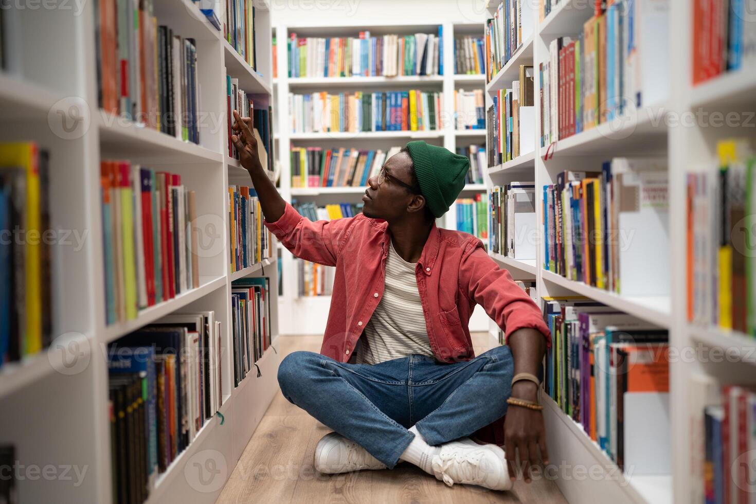 Young African American guy sits on floor in library between bookshelves, student searching for book photo