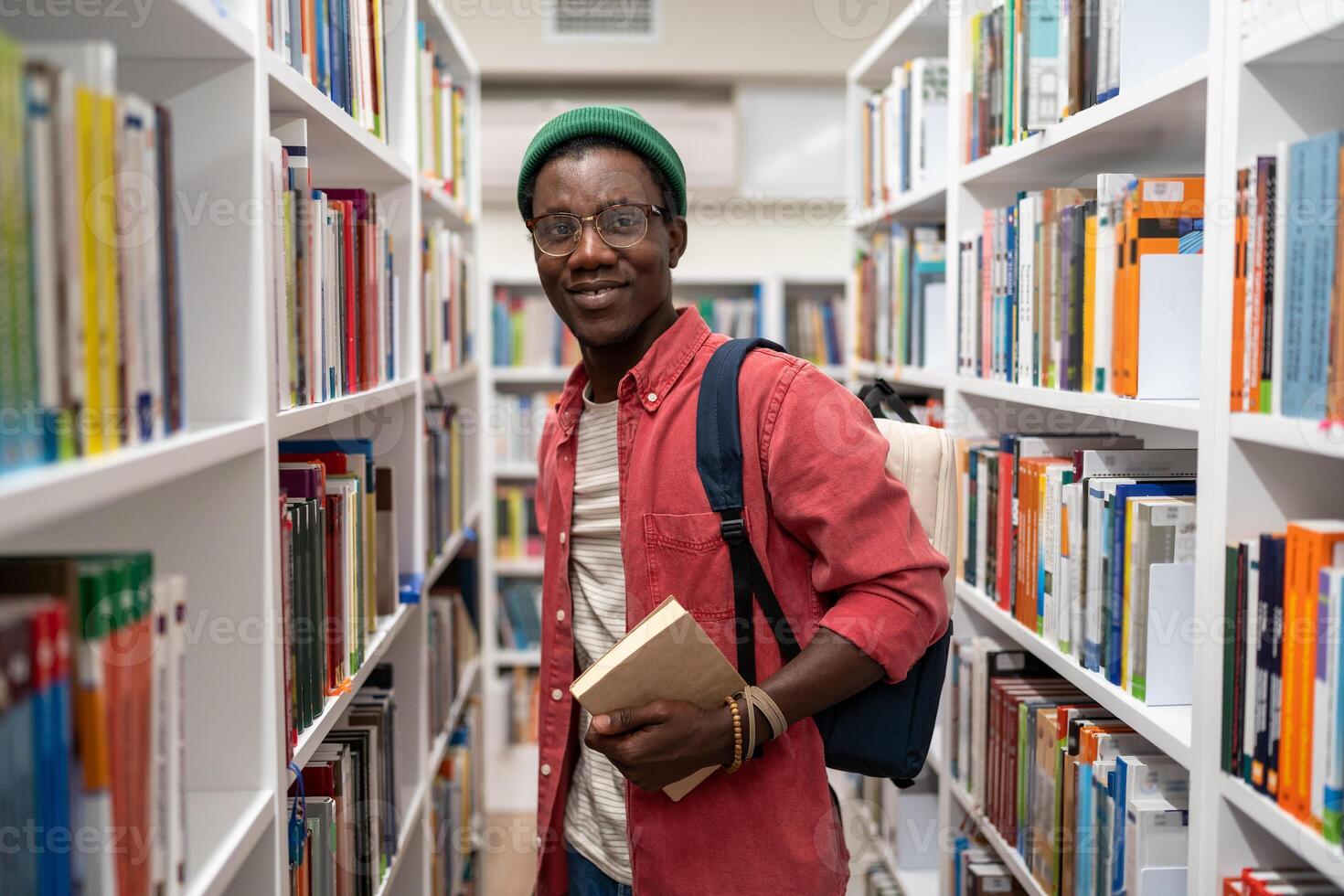 Smiling african american man student holds book in university library between bookshelves. photo