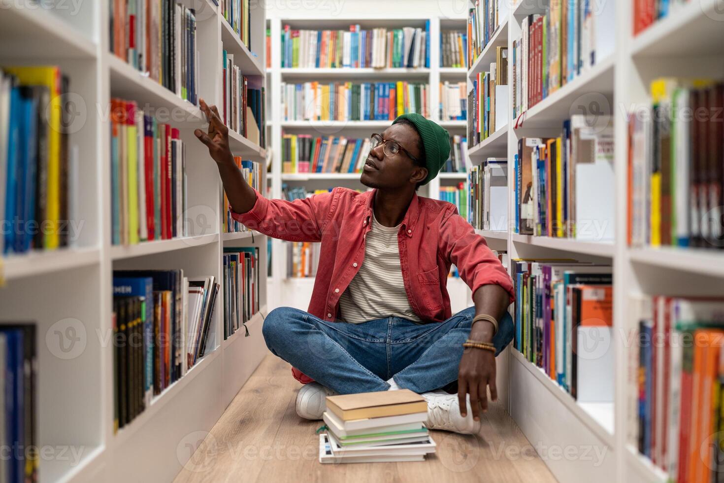 Young African American guy sits on floor in library between bookshelves, student searching for book photo