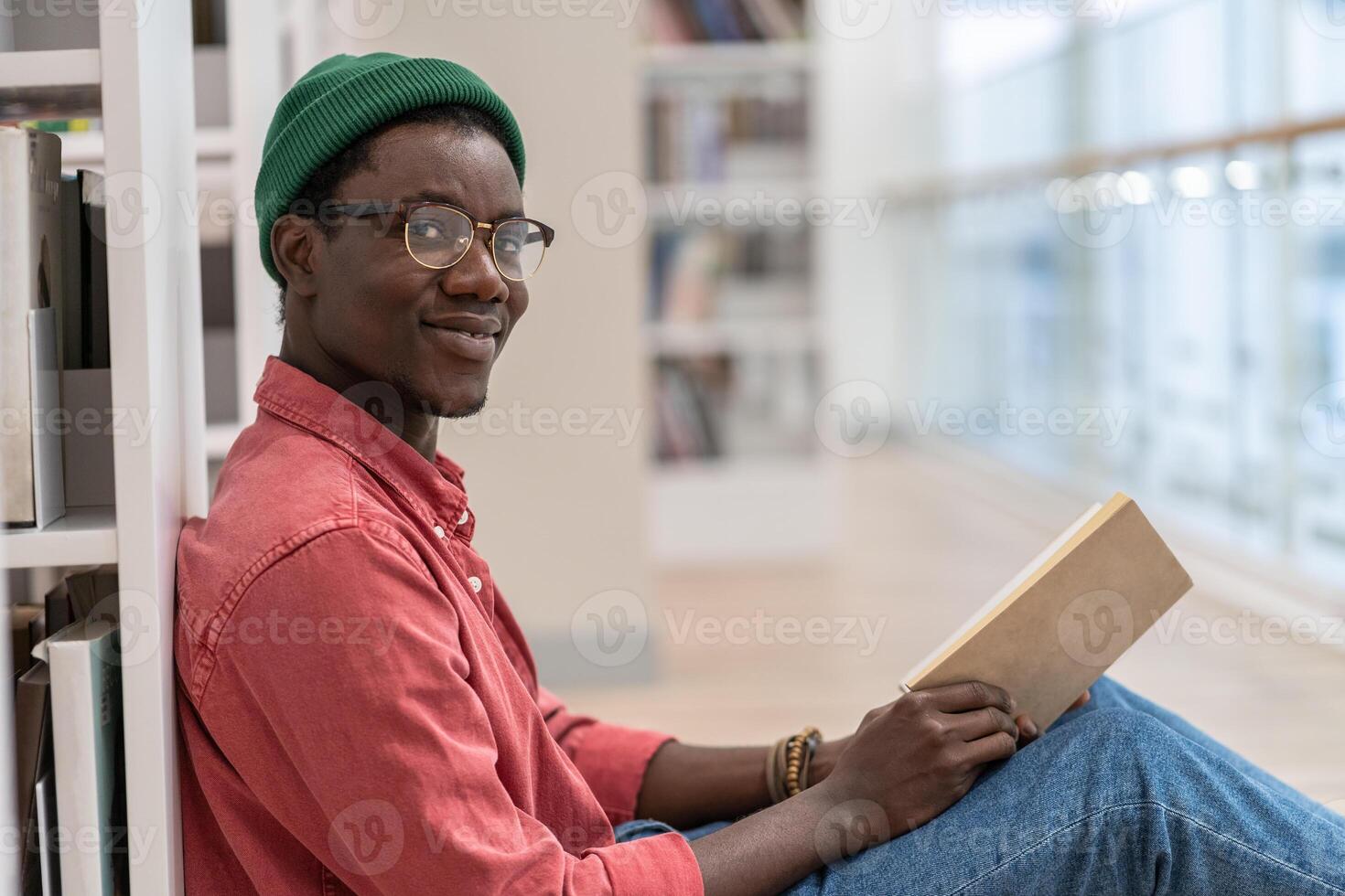 Black student man library visitor sitting on floor near bookcase with book in hands, looks at camera photo