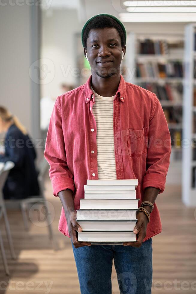 Portrait of happy African American student guy with pile of books in library photo