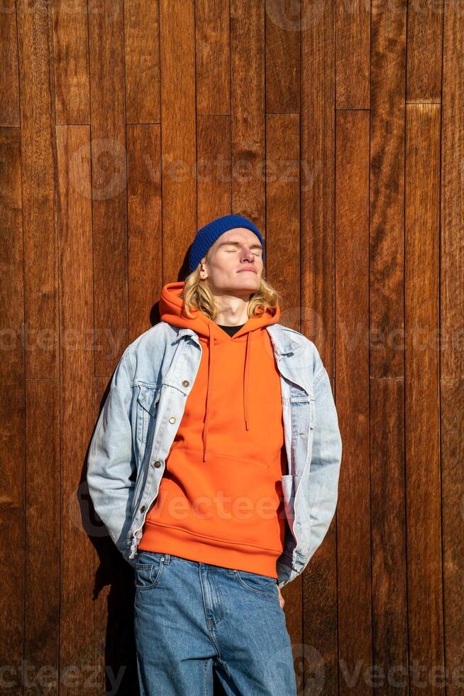 Relaxed skinny long-haired male with eyes closed leaning on wooden wall outdoors in bright sunny day photo