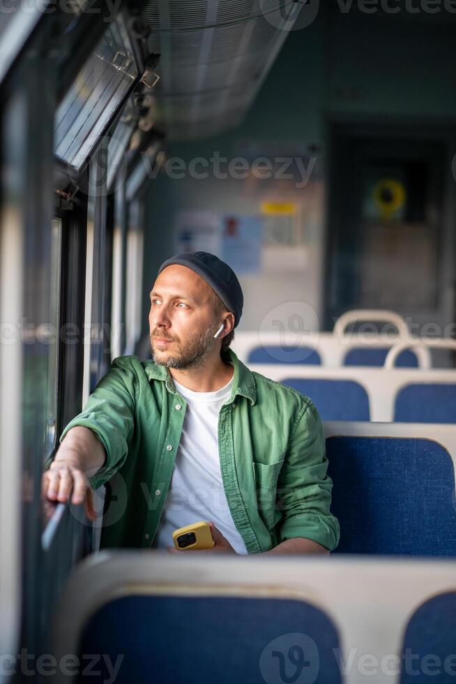 Thoughtful man listen to music in wireless earphones, phone in hand, gazing out train window photo